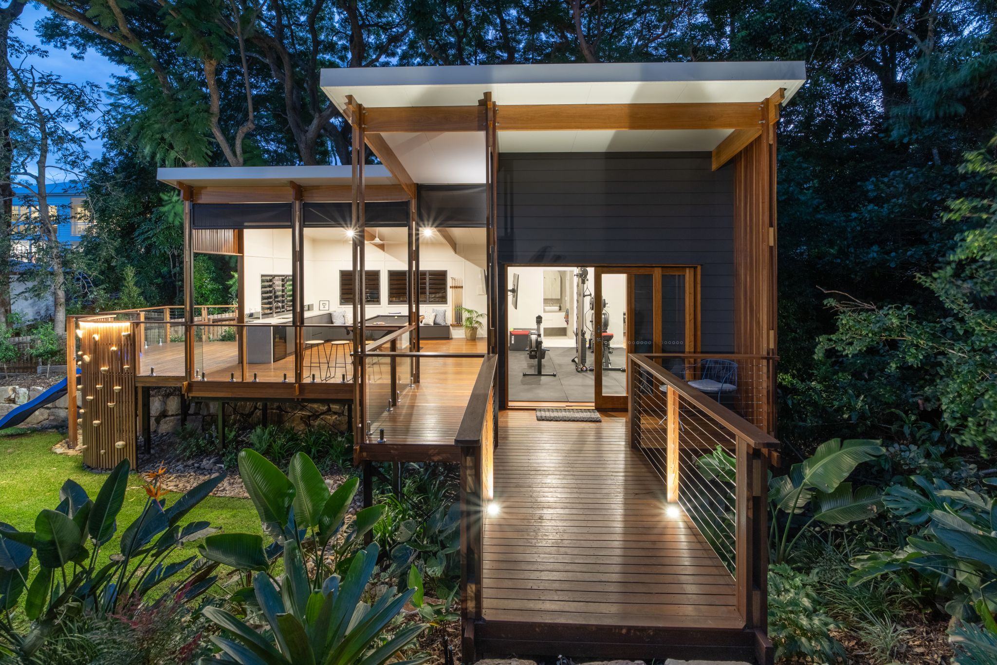 A modern house during twilight, featuring a wooden deck with EnduroShield-treated glass railings, reflecting the warm glow of the interior lights. The house is surrounded by lush greenery, and the deck leads to an open-concept interior with visible gym equipment. The structure showcases a combination of dark grey siding, natural wood posts, and a white flat roof, harmoniously blending with the natural environment.