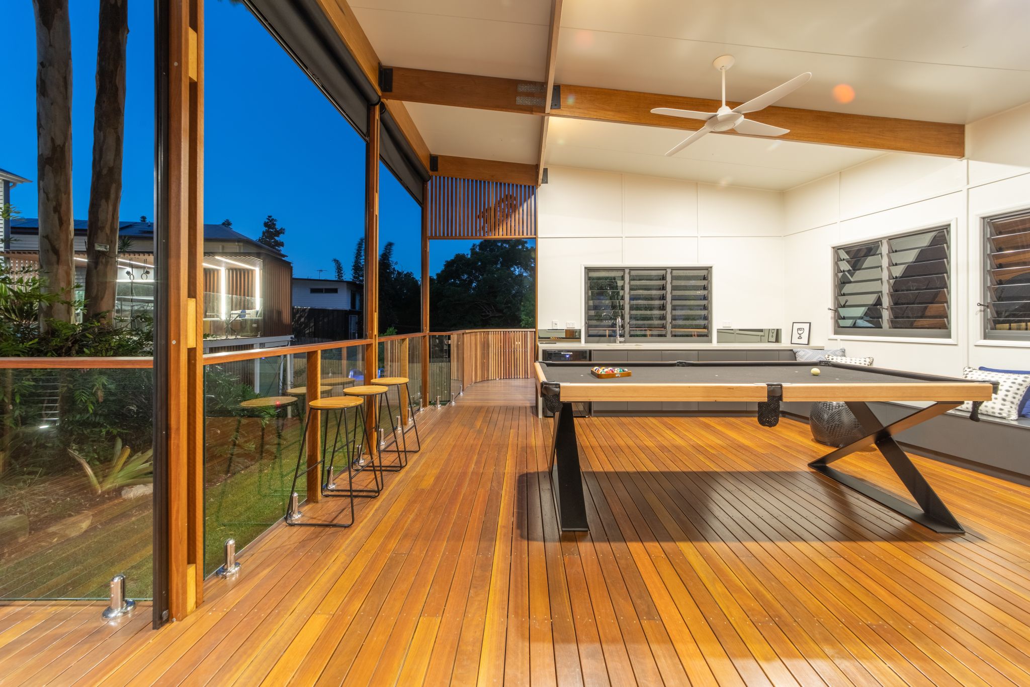 An expansive wooden deck during early evening, featuring a high-gloss EnduroShield finish that enhances the wood's natural grain and provides protection. The deck is furnished with bar stools and boasts a large, stylish black ping-pong table. Glass balustrades with sleek metal posts offer an unobstructed view of the neighboring area. The indoor space is visible through large windows and includes a modern kitchen and living area, unified by warm wood tones and a clean white ceiling with a ceiling fan.