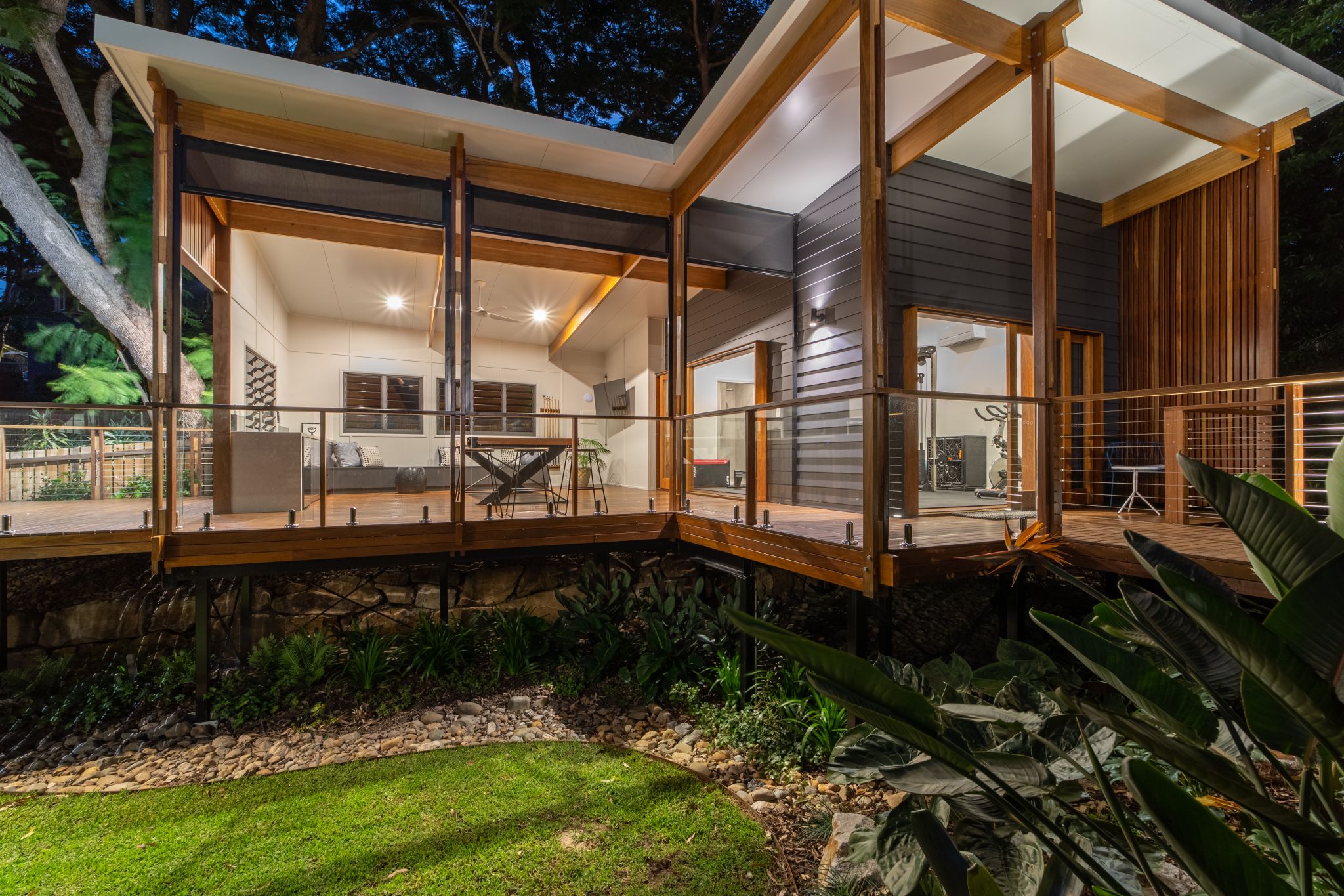 A modern house during twilight, featuring a wooden deck with EnduroShield-treated glass railings, reflecting the warm glow of the interior lights. The house is surrounded by lush greenery, and the deck leads to an open-concept interior with visible gym equipment. The structure showcases a combination of dark grey siding, natural wood posts, and a white flat roof, harmoniously blending with the natural environment.