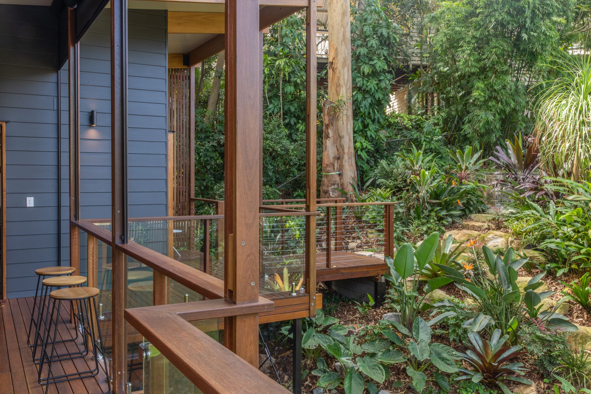 A tranquil outdoor deck area of a modern home, bordered by EnduroShield-protected glass railings that merge seamlessly with the natural surroundings. The wooden deck, adorned with stylish bar stools, steps down to a lush garden filled with a variety of tropical plants and foliage. The house's dark grey siding contrasts with the warm tones of the wooden beams and deck, while the garden path suggests a serene walk amidst greenery.