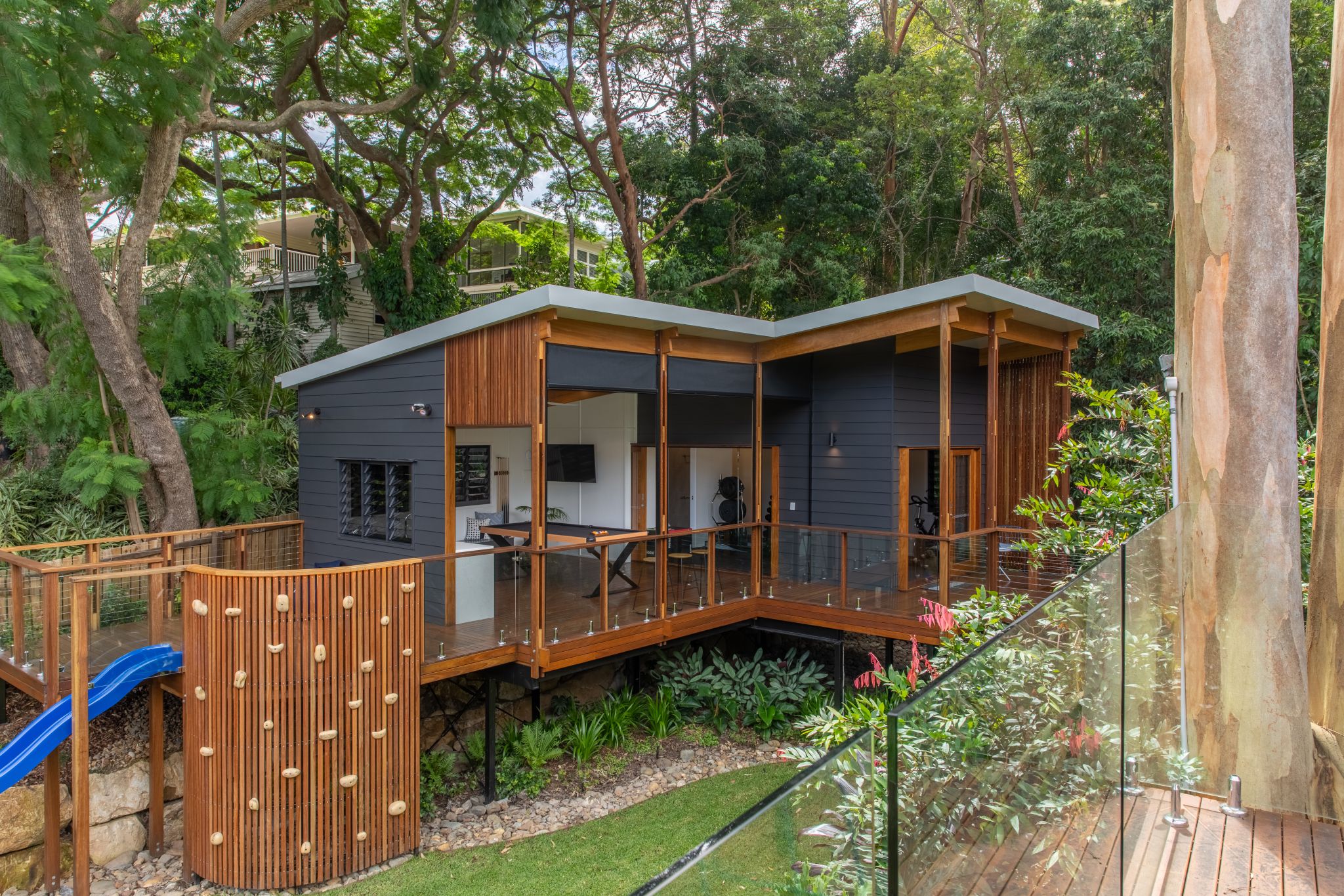 A modern family home set in a lush garden with a spacious backyard featuring a vibrant green lawn. The house is elevated on stilts, with a wooden deck that includes EnduroShield-coated glass railings, offering a clear view of the surrounding nature. A blue slide adds a playful touch, suggesting a child-friendly environment. The architecture displays a mix of dark grey and natural wood finishes, with large windows that allow for an indoor-outdoor living experience. The garden is rich with mature trees and tropical plants, enhancing the home's serene and private atmosphere.